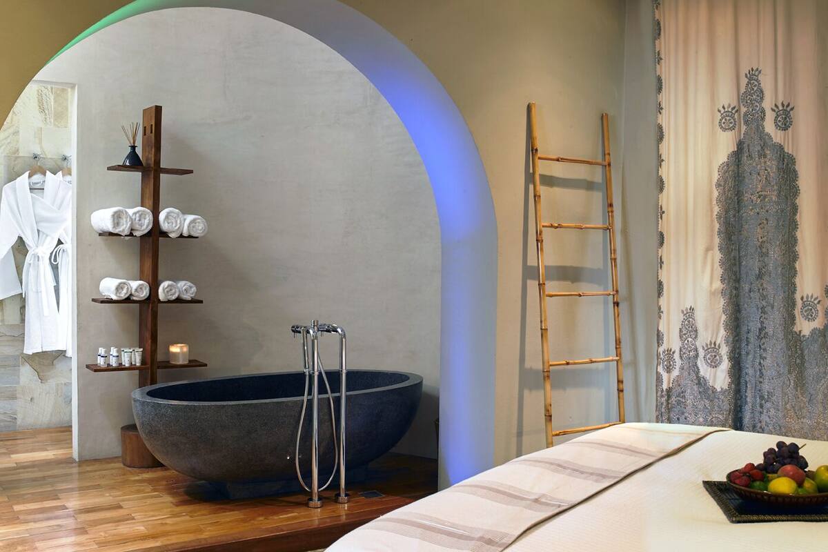 Een moderne badkamer met een groot stenen bad, een houten plank met handdoeken, een bamboe ladder en een fruitschaal op de wastafel.