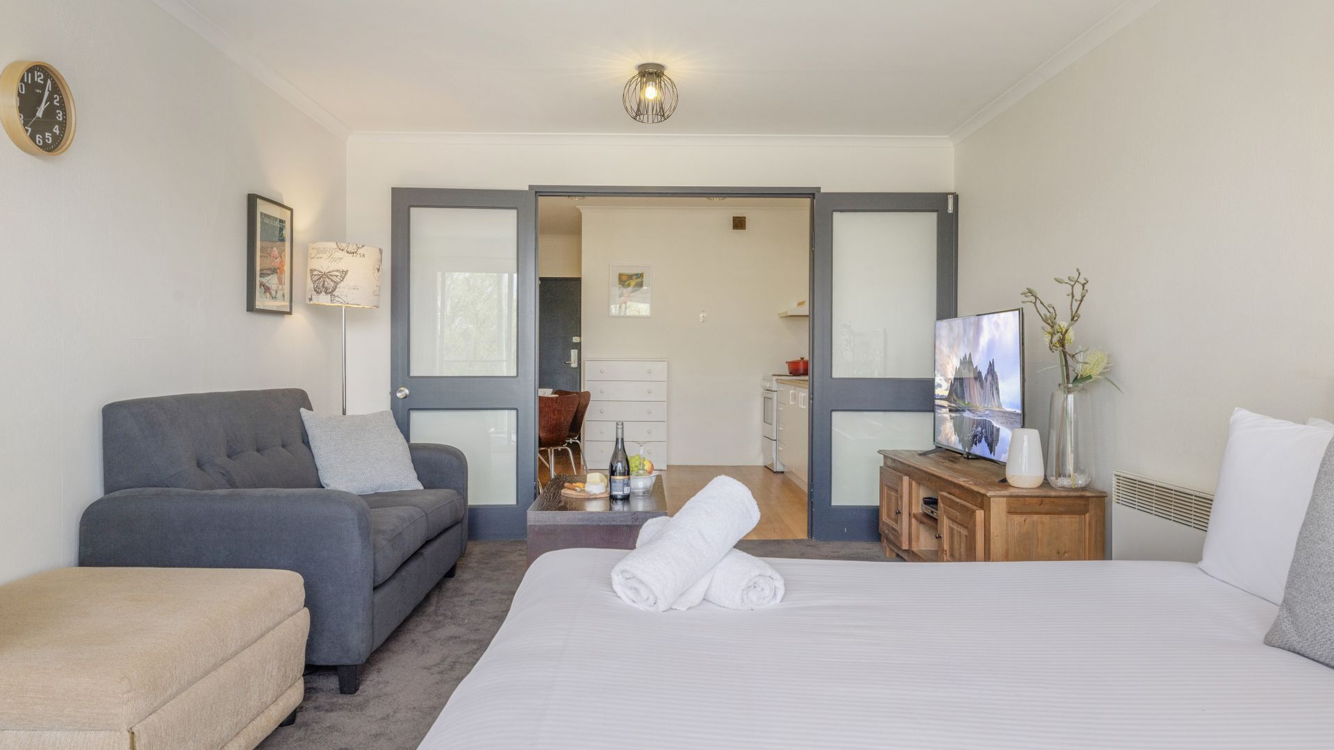 A modern hotel room with a grey sofa, a bed with white linens, a wooden side table, and a television.