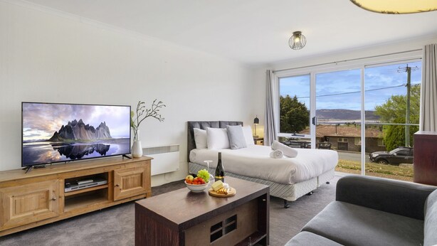 A modern hotel room with a bed, a television, a wooden cabinet, and a dining table with fruit and cheese.