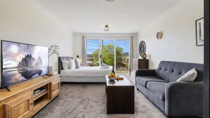 A modern living room with a large flat-screen TV, a grey sofa, a wooden coffee table, and a white sofa with a view of the outdoors.