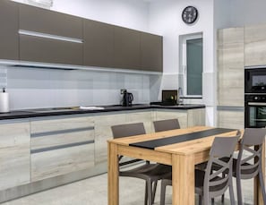 A modern kitchen with a wooden dining table and chairs, a clock on the wall, and a window.