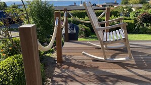 Un rivestimento in legno con una sedia a dondolo e una ringhiera in corda, con vista su un giardino e uno specchio d'acqua.