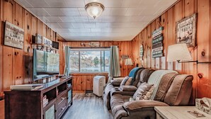 Ein gemütliches Blockhaushaus-Interieur mit Holzausstattung, einem Fernseher, einer Couch und einem Fenster mit Blick auf Bäume.