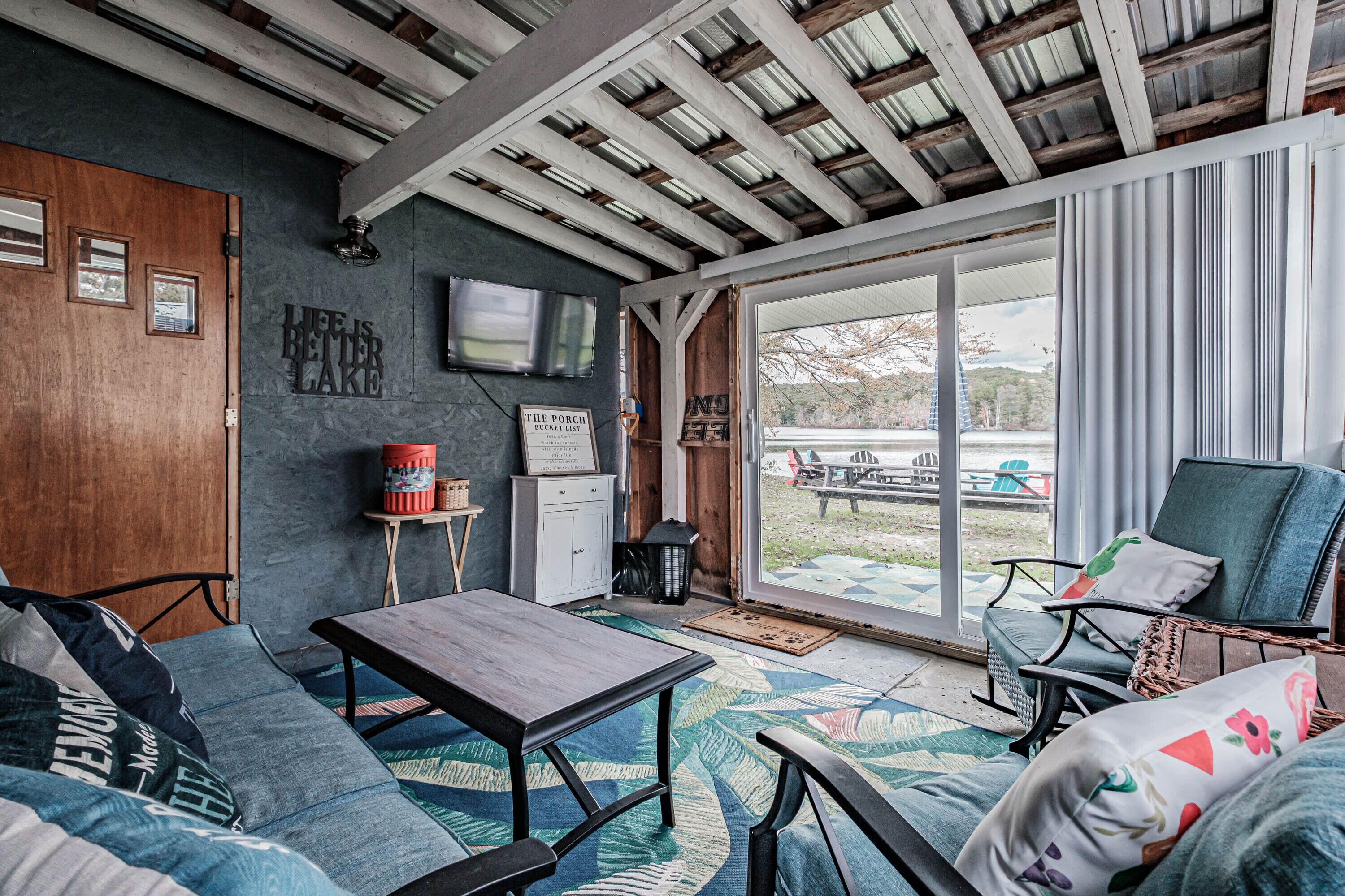 A cozy living room with a wooden ceiling, a sofa, a table, and a view of a backyard through large sliding glass doors.