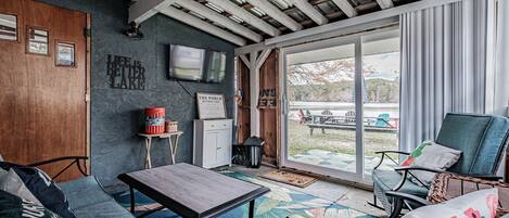 A cozy living room with a wooden ceiling, a sofa, a table, and a view of a backyard through large sliding glass doors.
