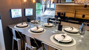 Uma área de refeições moderna com uma mesa comprida posta para seis pessoas, com luminárias pendentes geométricas e vista para um quarto com cama e janela.