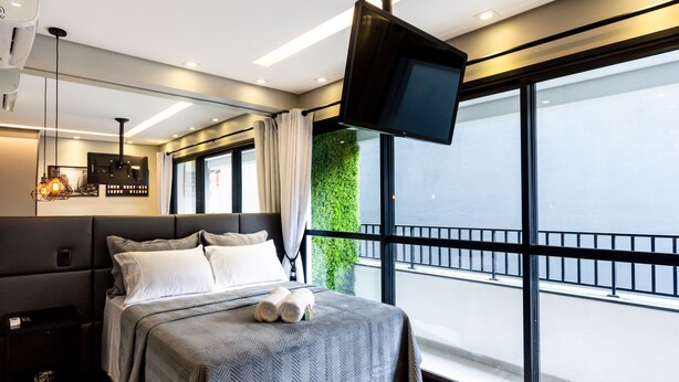 Quarto de hotel moderno com uma cama grande, televisão de tela plana na parede e uma varanda com vista para a cidade.
