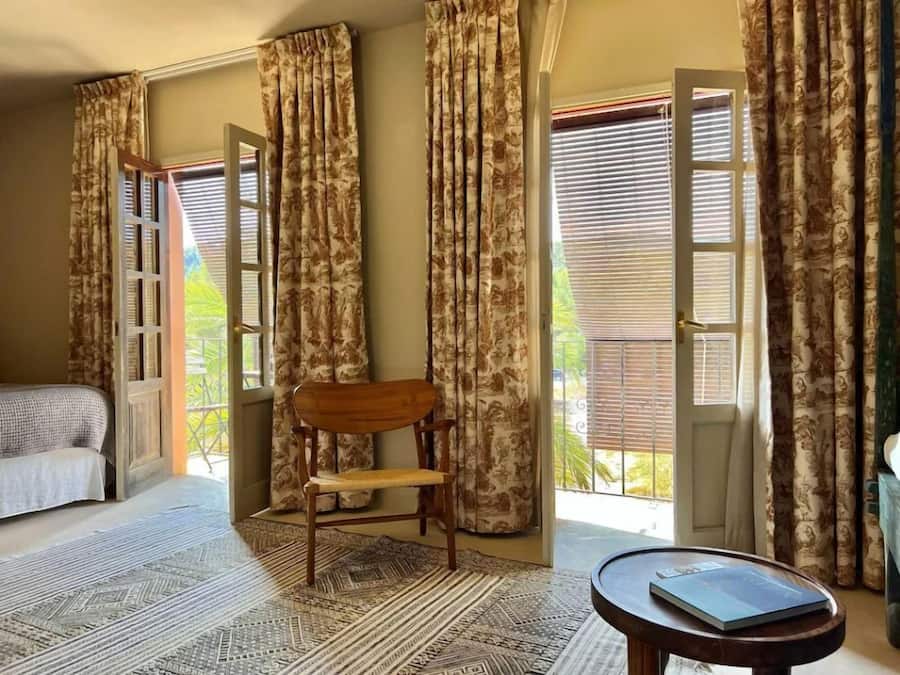 Una habitación con una cama, una silla de madera, una mesita y ventanas grandes con cortinas.