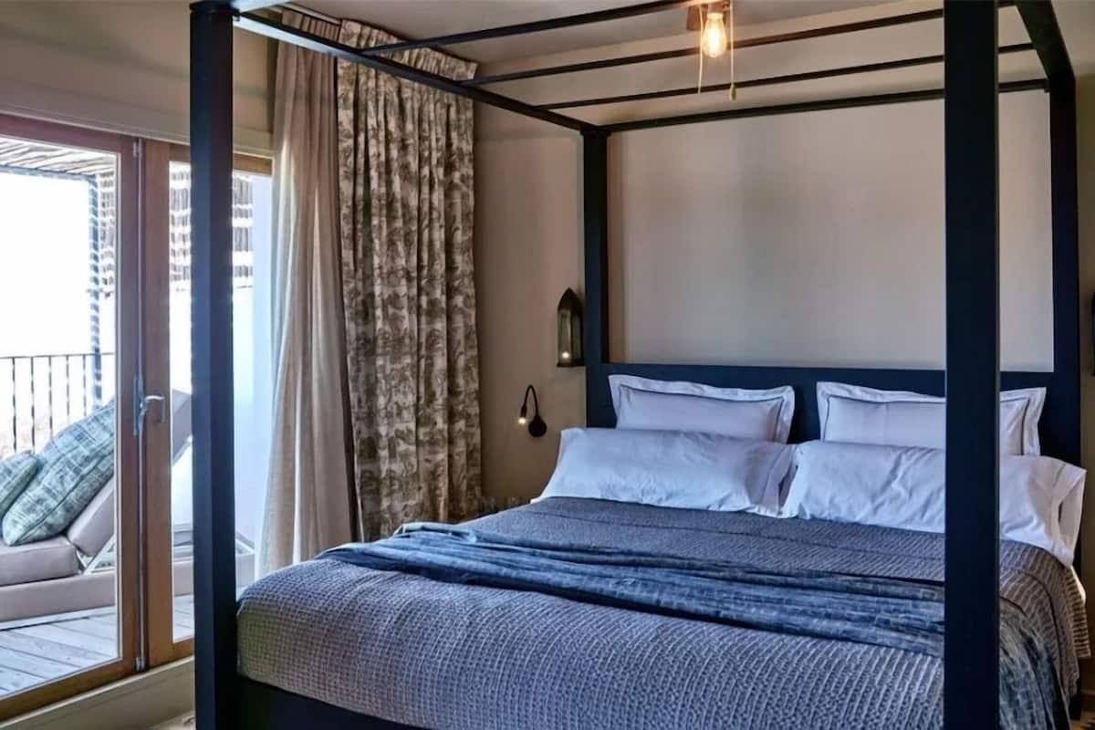 Un lit à baldaquin avec du linge de lit blanc et une couverture bleue, un balcon avec un canapé et des rideaux, et un petit luminaire fixé au mur.