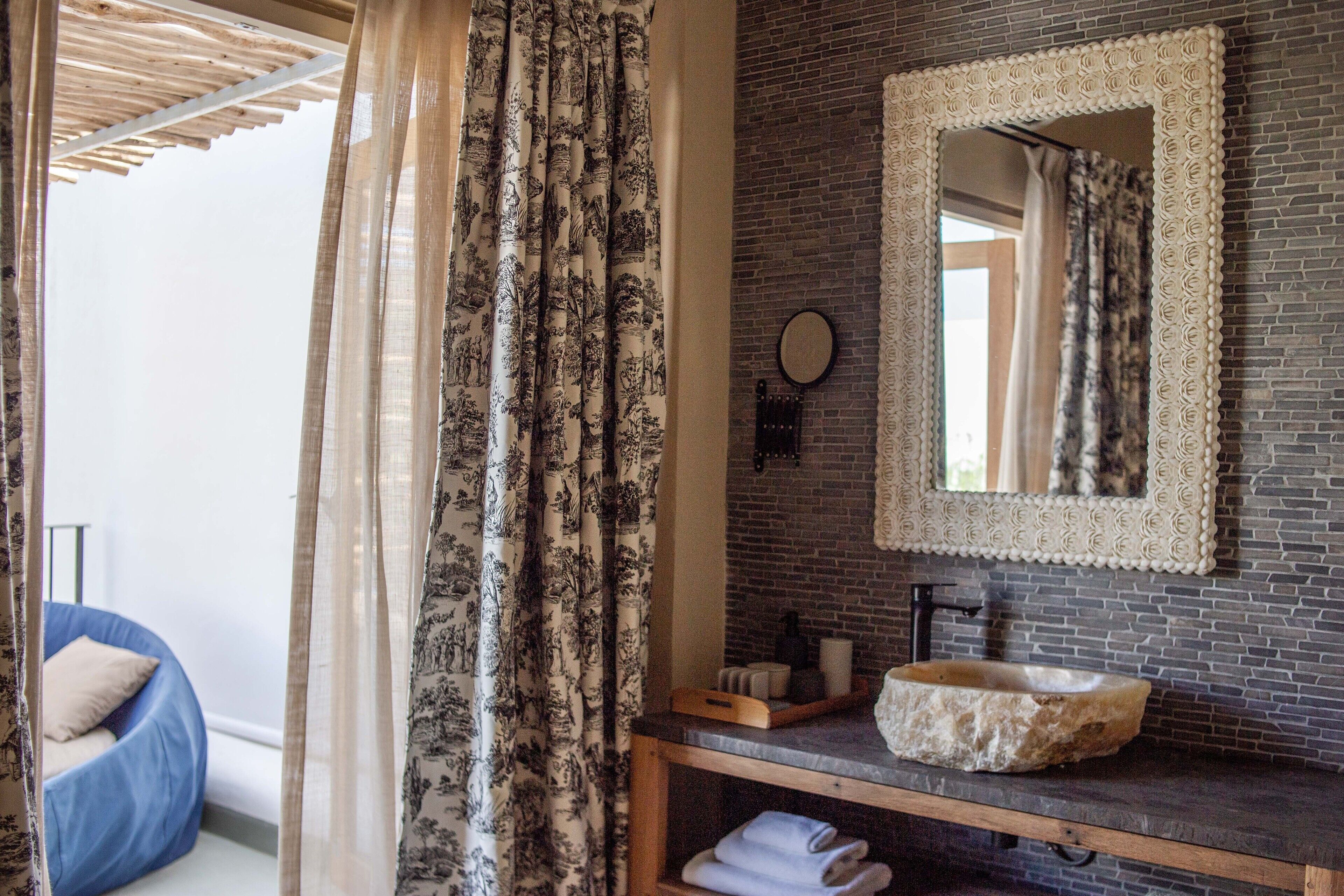 Een badkamer met een stenen wastafel, een spiegel met een sierlijk lijstwerk, en een blauwe kruis-kussen stoel op de achtergrond.