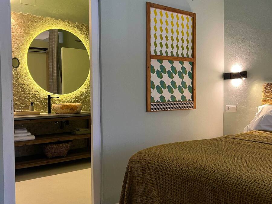 Un dormitorio con cama, un espejo redondo con marco texturizado, una lámpara de pared y un patrón geométrico enmarcado.