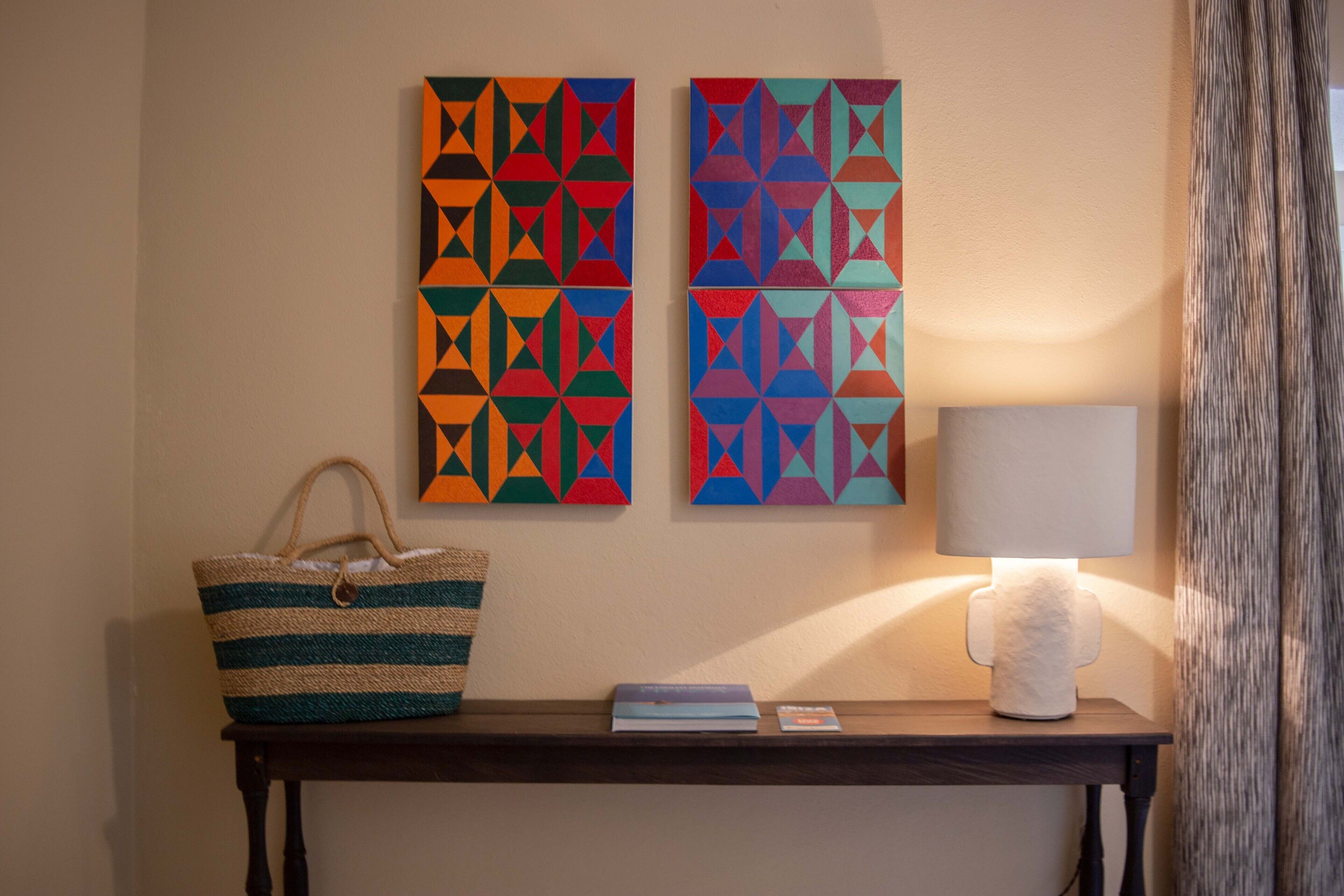 A wall with two colorful geometric paintings, a striped woven bag, a small table with a lamp, and a book.