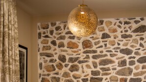 A room with a stone wall, a bed with patterned pillows, a wicker chair, a small round table, and a hanging light fixture. - Gare du Nord Hotel (Sant Joan de Labritja)