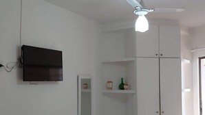 A room with a flat-screen TV mounted on the wall, a ceiling fan, a bed with a floral bedspread, and a wardrobe with a mirror. - Ap Banho de Mar (Ondina Salvador)