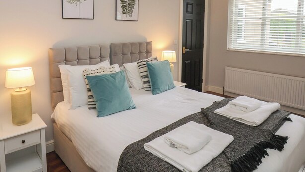 A bedroom with a large bed, two bedside lamps, a door, and a window with blinds. - The Pheasantry (Bridlington)
