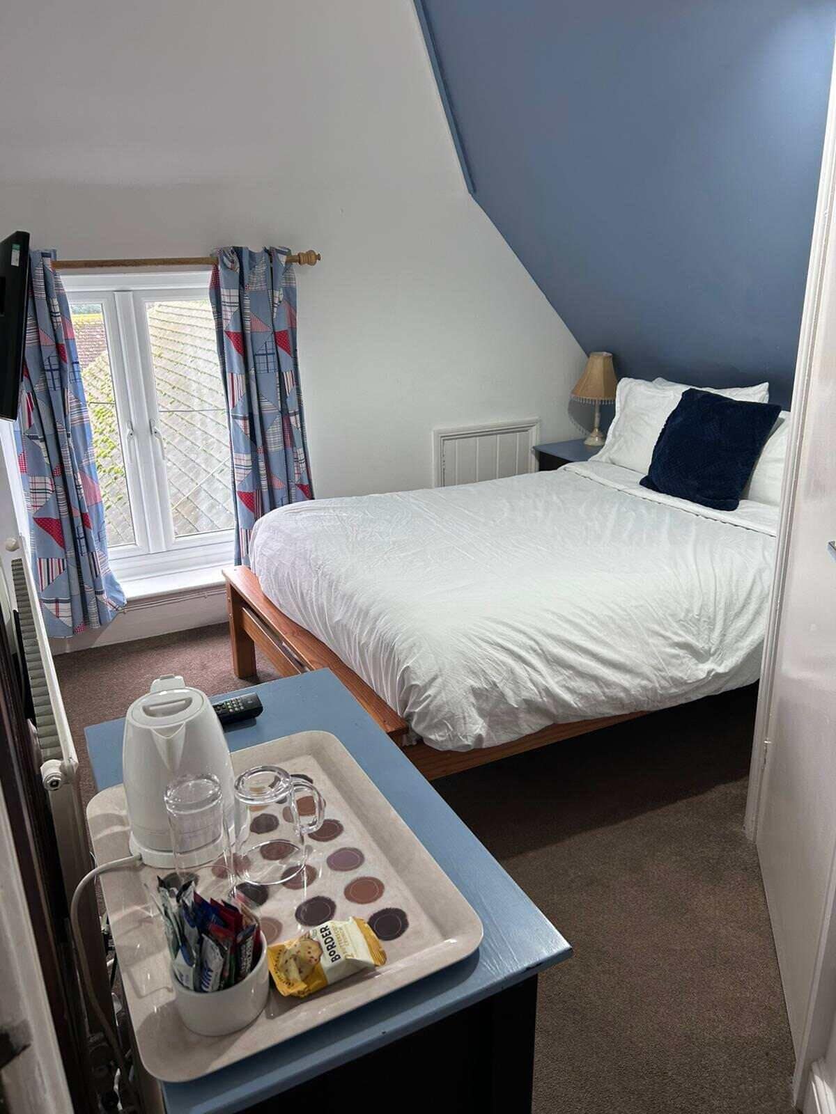 A bedroom with a bed, a bedside table with a tray containing a coffee pot, glasses, and snacks, a window with curtains, and a radiator.