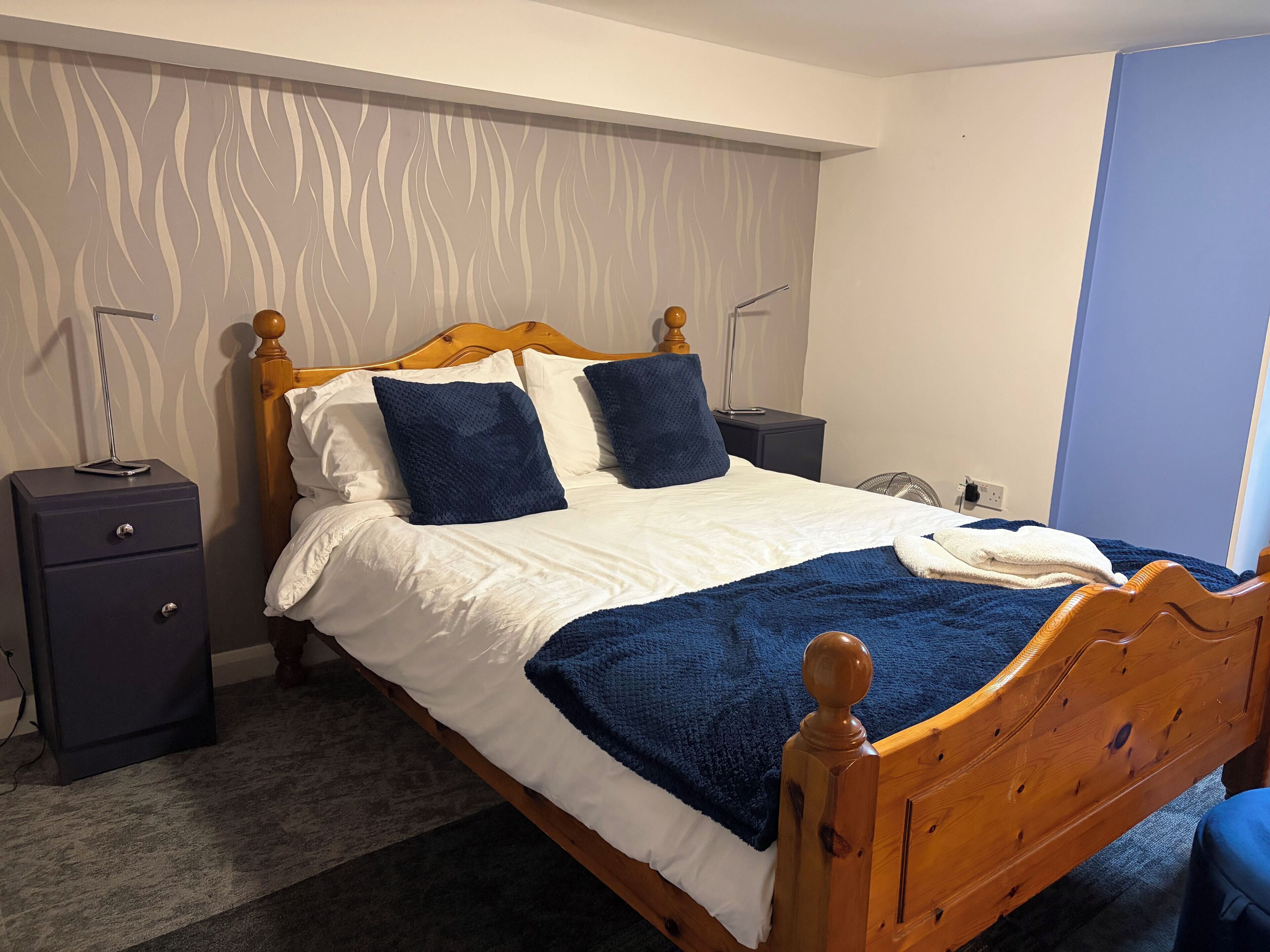 Una cama bien hecha con ropa de cama blanca y azul, un cabecero de madera y una mesita de noche oscura con una lámpara.