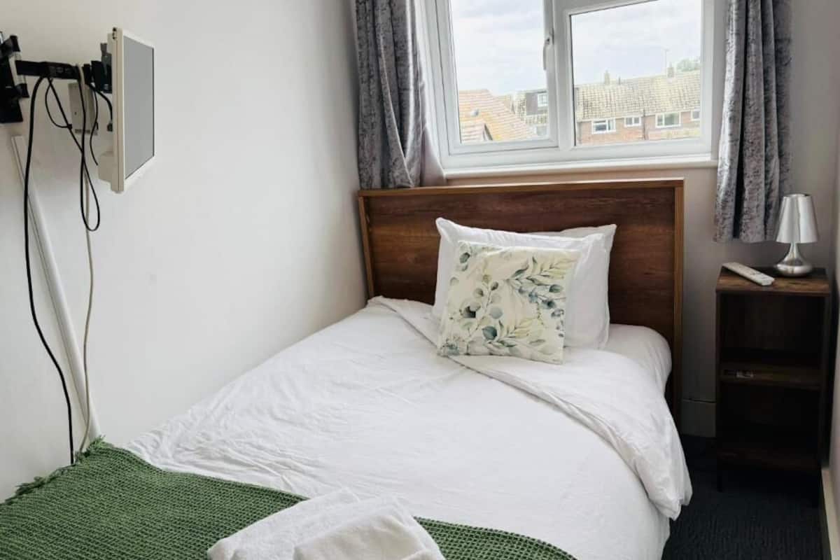 A single bed with a wooden headboard, a green throw blanket, a white pillow with a floral pattern, a bedside table with a lamp, and a window with curtains.