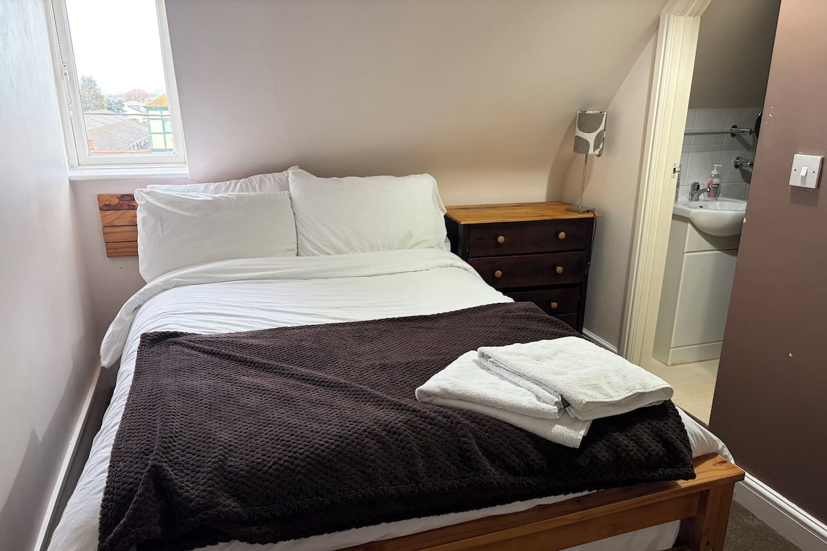 A neatly made bed with white linens, a dark brown blanket, and folded white towels. A wooden nightstand with a drawer is beside the bed. A window with a view of buildings is visible.