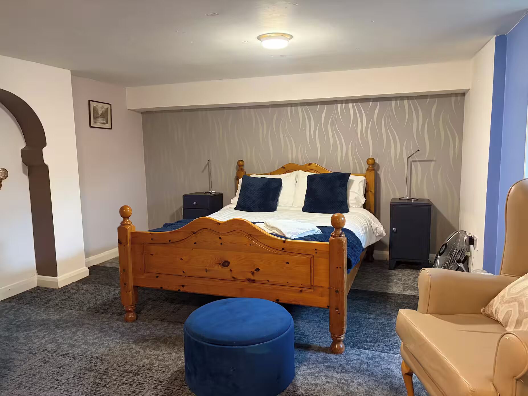 A bedroom with a wooden bed, a blue ottoman, a chair, and a nightstand with a lamp.