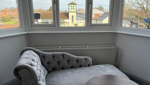 A room with a grey upholstered bench and a round table, large windows with a view of buildings and a clock tower. - Captain Howey Hotel (New Romney)