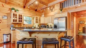 Interior de una cabaña de madera con cocina que incluye una isla grande, taburetes de bar y un refrigerador.