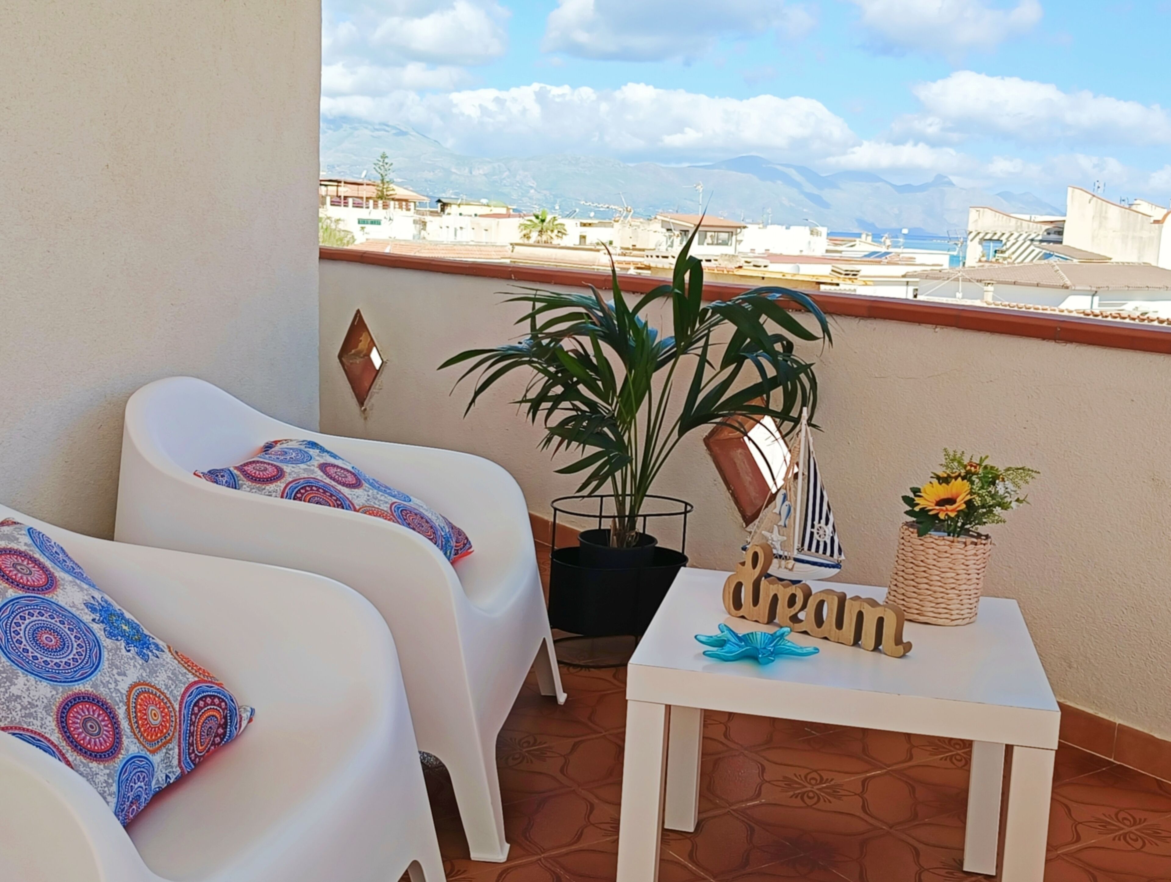 A balcony with two white chairs, a potted plant, a small table with a sunflower vase, and a decorative sailboat model.