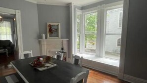 Un coin repas avec une table noire et des chaises blanches, un lustre et une fenêtre avec des volets blancs.
