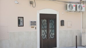 Fachada de un edificio con una puerta marrón, dos unidades de aire acondicionado, una cámara de seguridad y un letrero que indica una habitación llamada "B&B Kapa Villa Room's".