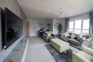 Ein modernes Wohnzimmer mit Flachbildfernseher, grauen Sofas, einem Couchtisch und einem Essbereich im Hintergrund.