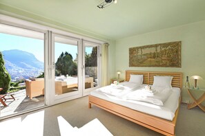 A bedroom with a large bed, a painting, and a view of a mountainous landscape through a sliding door.