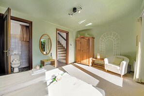 A spacious bedroom with a large bed, a wooden wardrobe, a white armchair, and a staircase leading to another level.