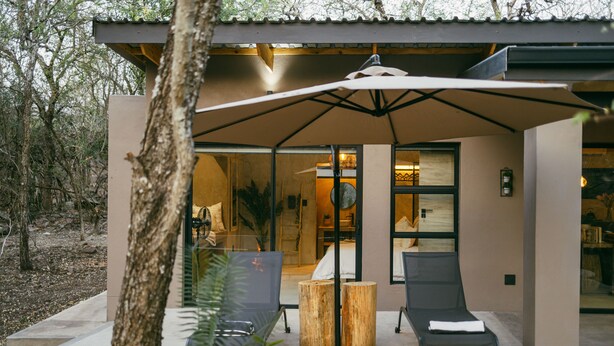 Un área de estar al aire libre de estilo moderno con una mesa, sillas y un taburete de madera, además de un gran paraguas que proporciona sombra.