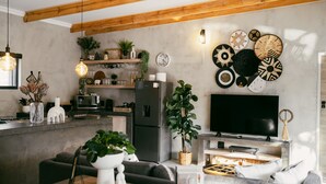 Una sala moderna con sofá, mesa de centro y televisor. Cuenta con una zona de cocina con refrigerador, estantes y plantas.