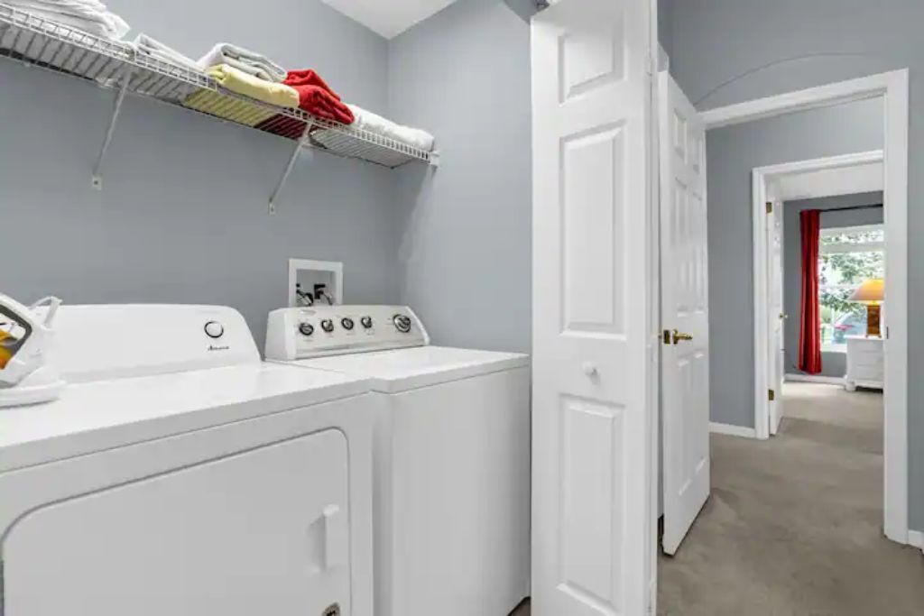 A laundry room with a front-loading washer and dryer, a drying rack above, and a door leading to another room.