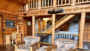 Interior de una cabaña de troncos con dos sillones tapizados, una escalera de madera y un tapiz con motivos forestales.