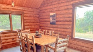 Comedor de una cabaña de madera con una mesa grande y sillas, un lámpara colgante y un cuadro en la pared.