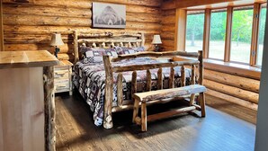 Un dormitorio de cabaña de troncos con una cama de madera, dos lámparas de noche, un cuadro en la pared y un ventanal con vistas a la vegetación.
