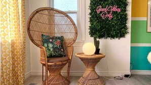 Une chaise en osier avec un coussin, une petite table ronde avec une lampe et une plante fixée au mur, accompagnée d'une pancarte indiquant « Seules les bonnes ondes sont les bienvenues ».