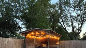Ein Garten mit Schwimmbad, Holzterrasse und überdachter Sitzgelegenheit mit Lichterkette.