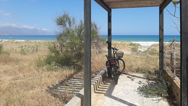 Un espacio al aire libre cubierto con una bicicleta estacionada bajo una estructura de madera, con vista a una playa de aguas azules cristalinas y montañas a lo lejos.