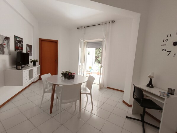 Una habitación moderna y bien iluminada con una mesa de comedor, sillas, un televisor sobre un mueble blanco y un escritorio pequeño con una lámpara.