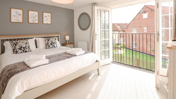 A bedroom with a bed, a bedside table, a lamp, a large window, and a view of a brick building. - The Mill Stones (Norwich)