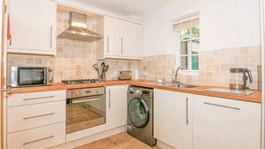 A modern kitchen with white cabinets, a built-in oven, microwave, dishwasher, and a window with blinds. - The Mill Stones (Norwich)