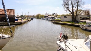 A canal with boats docked on both sides, a small building with a red roof, and a tree in the background. - The Mill Stones (Norwich)