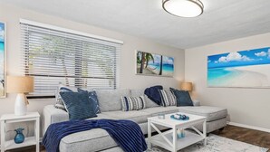 A living room with a grey sofa, white coffee table, and a blue throw blanket. There are two side tables with lamps and a painting of a beach scene. - Little Treasure House minutes from Beach & Mercato (Naples)
