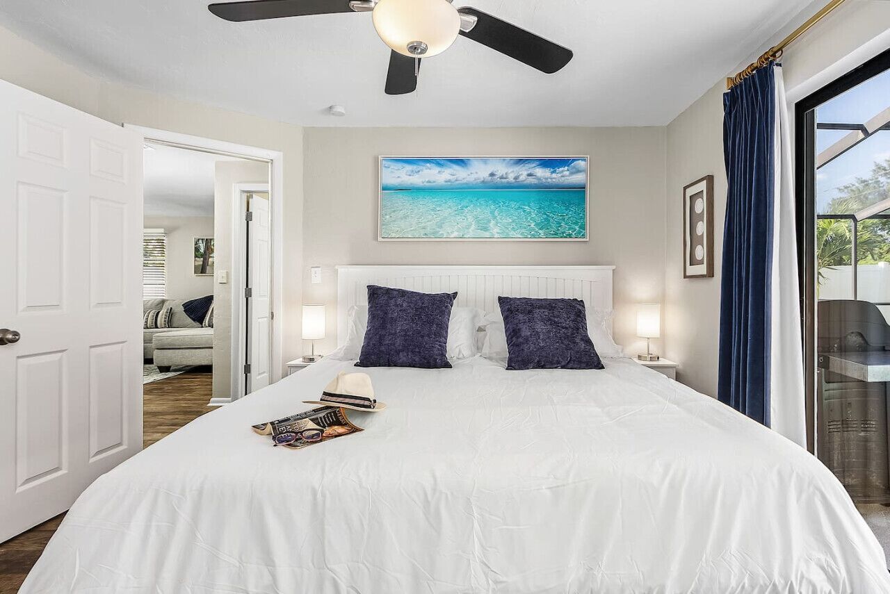 Ein Schlafzimmer mit einem Bett, einem Deckenventilator, einem Fenster mit Vorhängen und einem Bild einer Strandlandschaft.