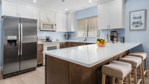 A modern kitchen with white cabinets, stainless steel appliances, a central island with stools, and a fruit bowl. - Tropical vacation home close to Vanderbilt Beach (Naples)
