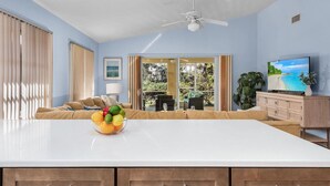 A modern living room with a large white island, a ceiling fan, and a television on a wooden cabinet. - Tropical vacation home close to Vanderbilt Beach (Naples)