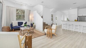 Un vaste espace de vie comprenant un canapé, une table basse et des fauteuils, ainsi qu’un escalier menant à une cuisine équipée de meubles blancs et d’un réfrigérateur.